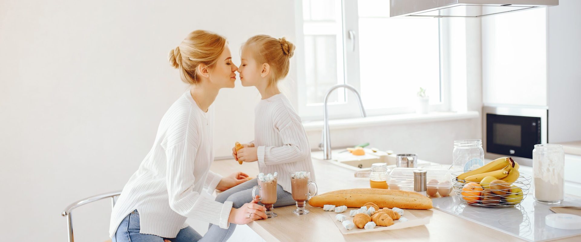 a beautiful young mother with light hair in white lace and blue jeans trousers sitting at home in the kitchen along with her little cute daughter and they drinking cappuccino